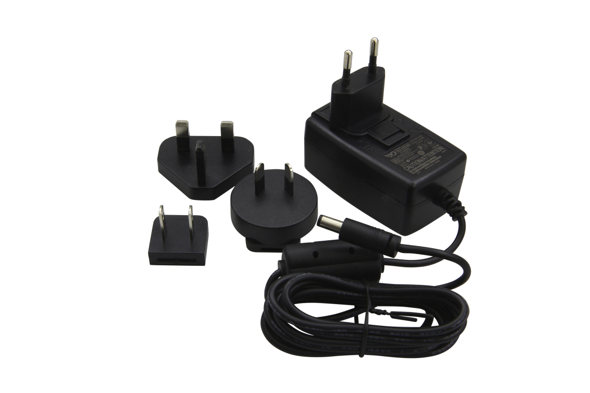 Schwarzes Kern-Steckernetzgerät mit EU-Stecker (2 runde Stifte). Daneben drei austauschbare Adapter: UK (3 rechteckige Stifte), US (2 flache Stifte) und CH (3 runde Stifte). Kabel zum Hohlstecker.