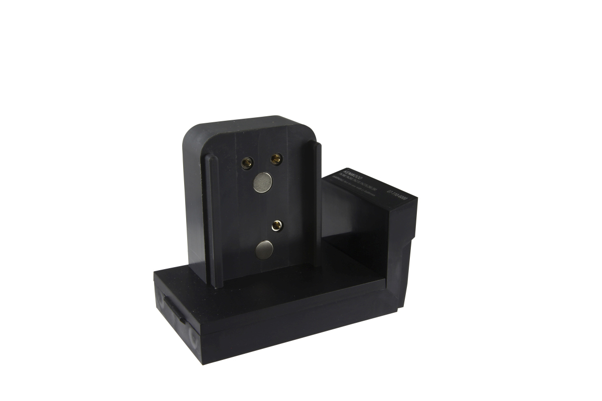 Adaptador negro « Cadex » para radios de la serie Kenwood con superficie blanca. En forma de L con parte trasera elevada, dos contactos magnéticos plateados y dos contactos de clavija dorados. En el lateral, «Kenwood» y «07-110-6000». Para KNB-14/KNB-15.
