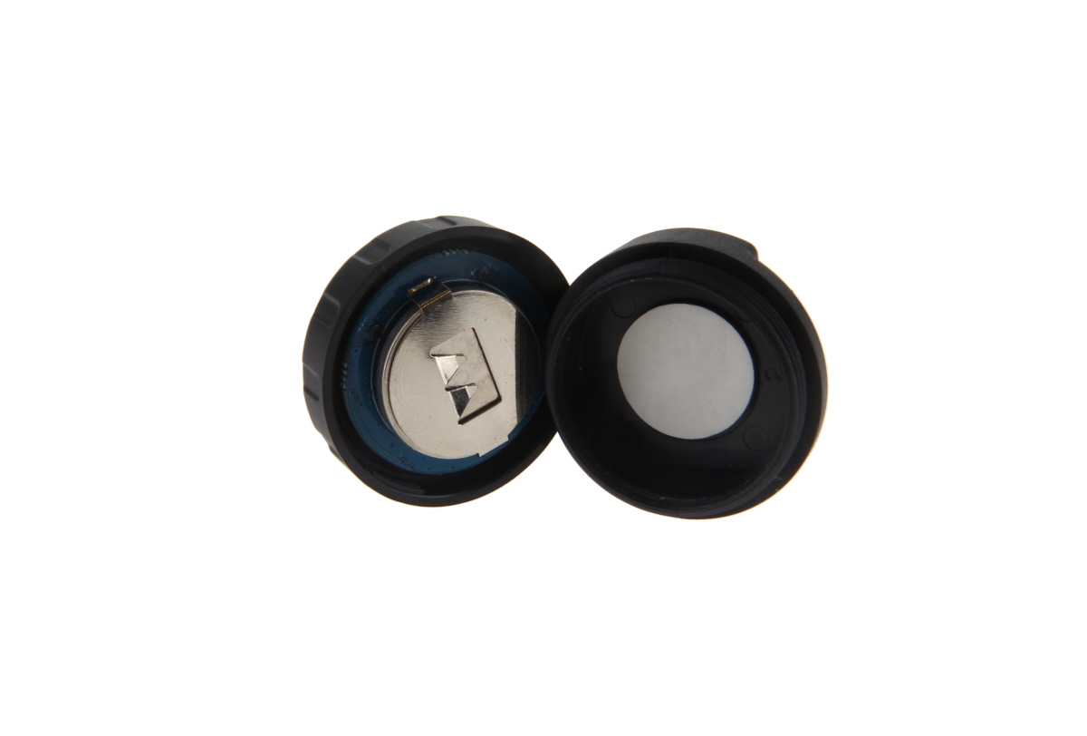 Alloggiamento nero rotondo aperto di un'unità PTT wireless con batteria a bottone CR2032 argentata inserita e contatti metallici. Coperchio accanto con guarnizione/punto di contatto bianco. Anello di tenuta blu (IP67).