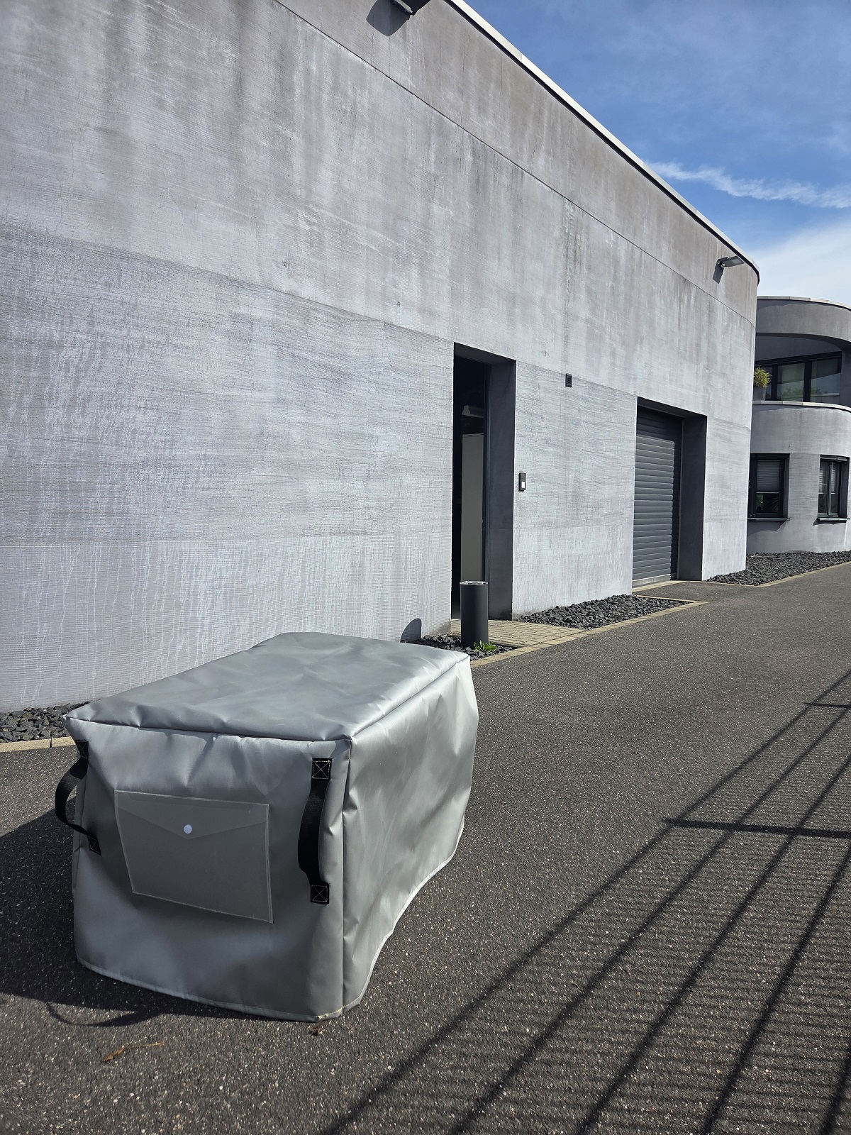 Copertura antincendio tipo H grigia rettangolare in tessuto di vetro HR con silicone ignifugo, sull'asfalto scuro davanti a un moderno edificio grigio con porte e garage. Con maniglie a passante e tasca portadocumenti.