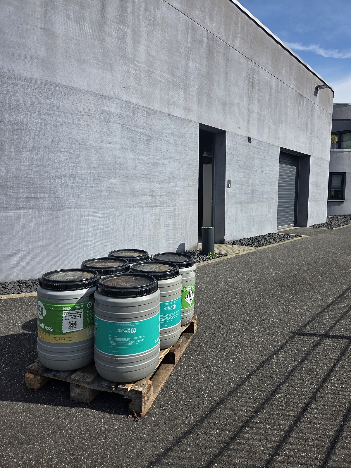 Sei fusti grigi con coperchi neri sono su un pallet di legno davanti a un moderno edificio grigio. Alcuni hanno etichette verdi; una mostra il simbolo di pericolo per batterie agli ioni di litio.