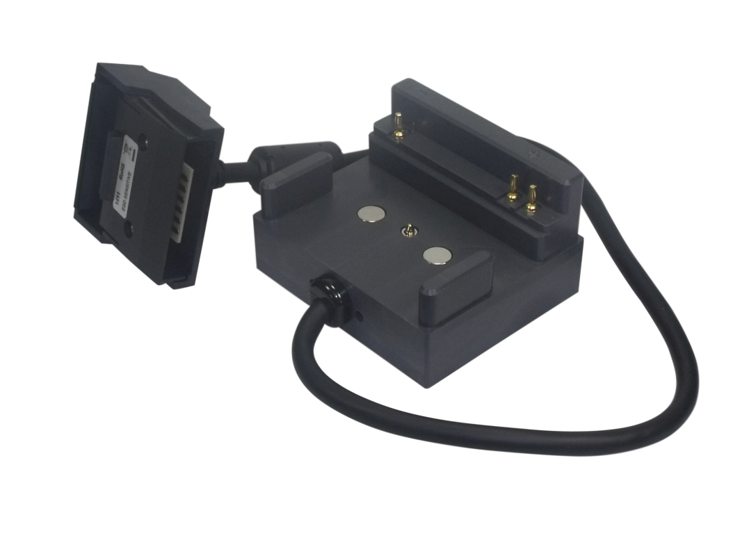 Adattatore nero " Cadex " per batterie serie M (PD4410) dell'Zoll. Due parti collegate: modulo rettangolare con connettore multiplo ed etichetta, nonché base con 2 contatti magnetici e 2 pin di ricarica dorati. Sono collegati tramite cavi.